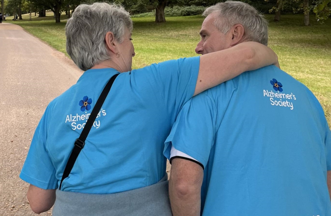 Chris and his wife on a Memory Walk. His wife has her arm around Chris, they are facing away from the camera wearing blue Alzheimer's Society Memory Walk t-shirts.