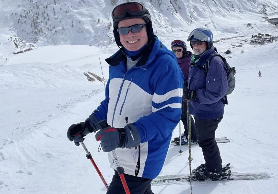 Chris at the top of a ski slope wearing skiing gear and skis. He is smiling at the camera with sunglasses on.