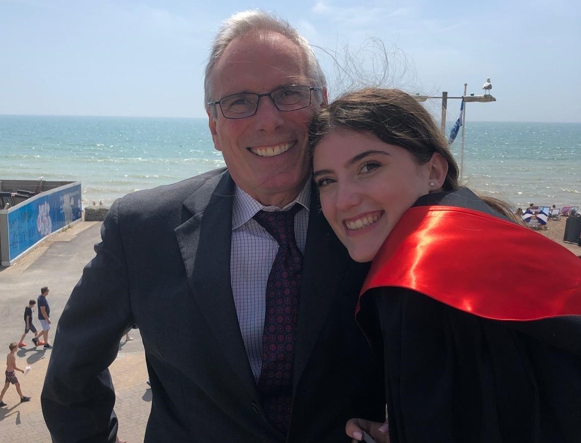 Lauren hugs her dad Chris, she is wearing a graduation gown and they are standing in front of the sea.