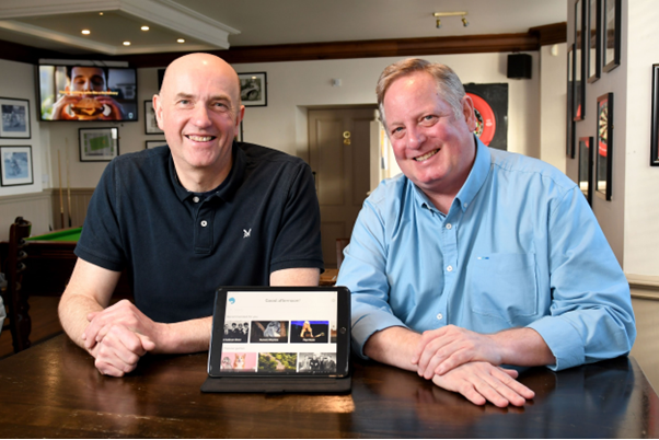An image of the two founders of Memory Lane Games, sat at a table with a tablet in front of them, showcasing the product. They are both smiling.