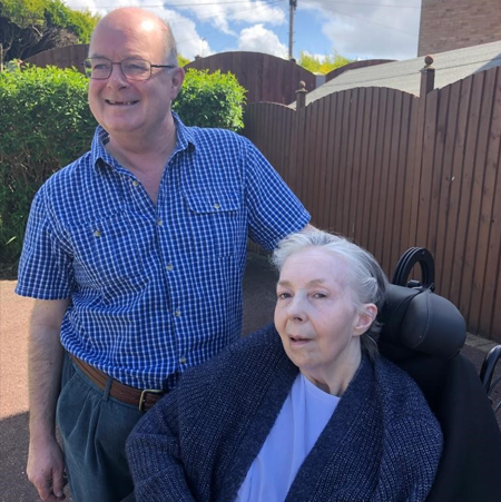 A smiling man stands next to his wife, who is in a wheelchair.