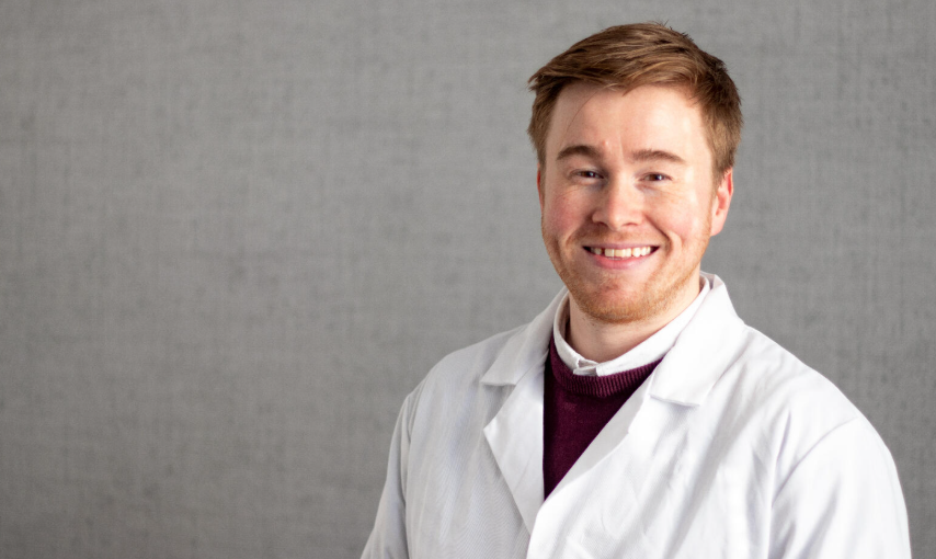 A young man in a white lab coat smiling.