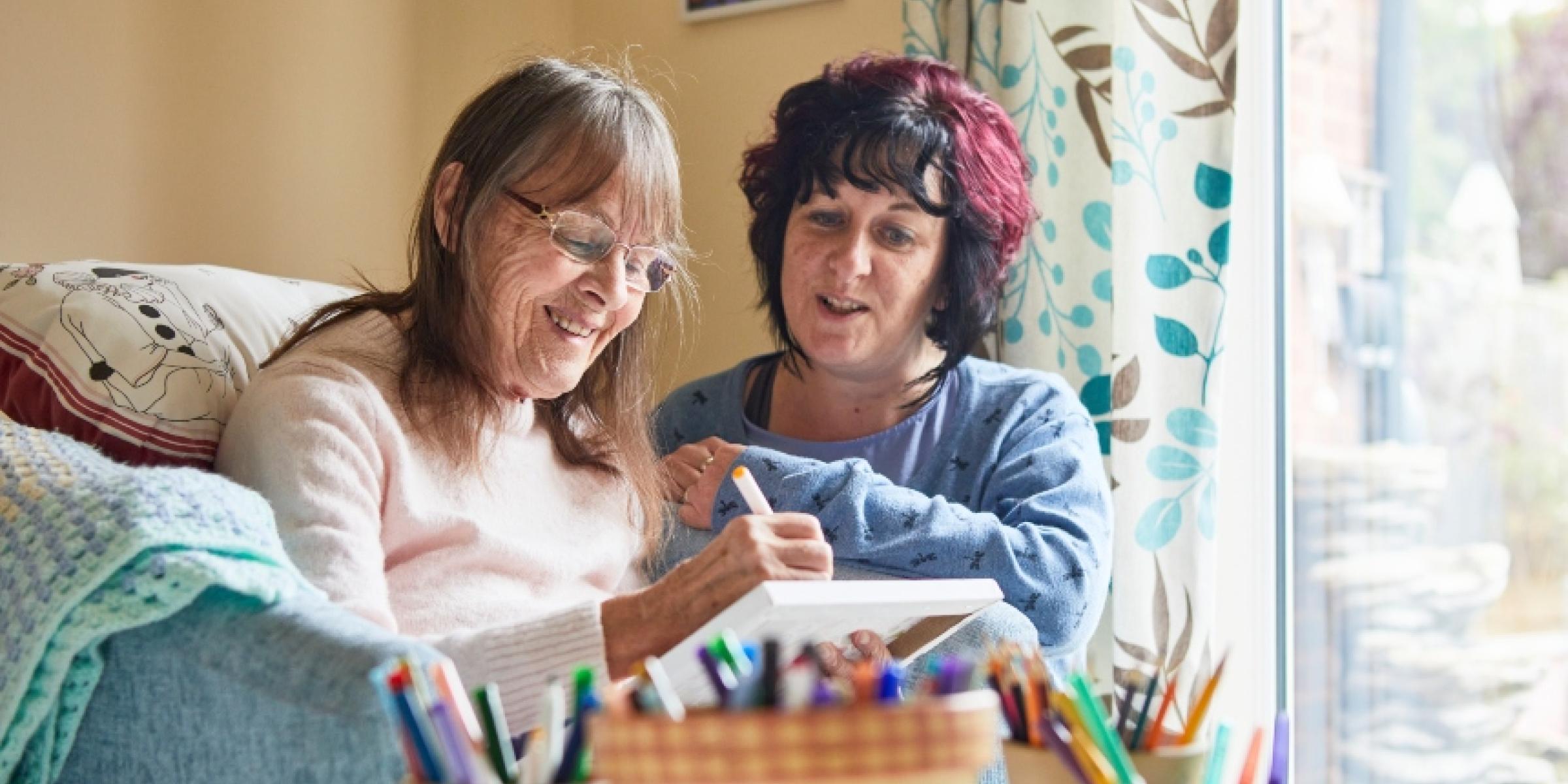 Two women sat side by side in their living room, using coloured pens to complete an activity