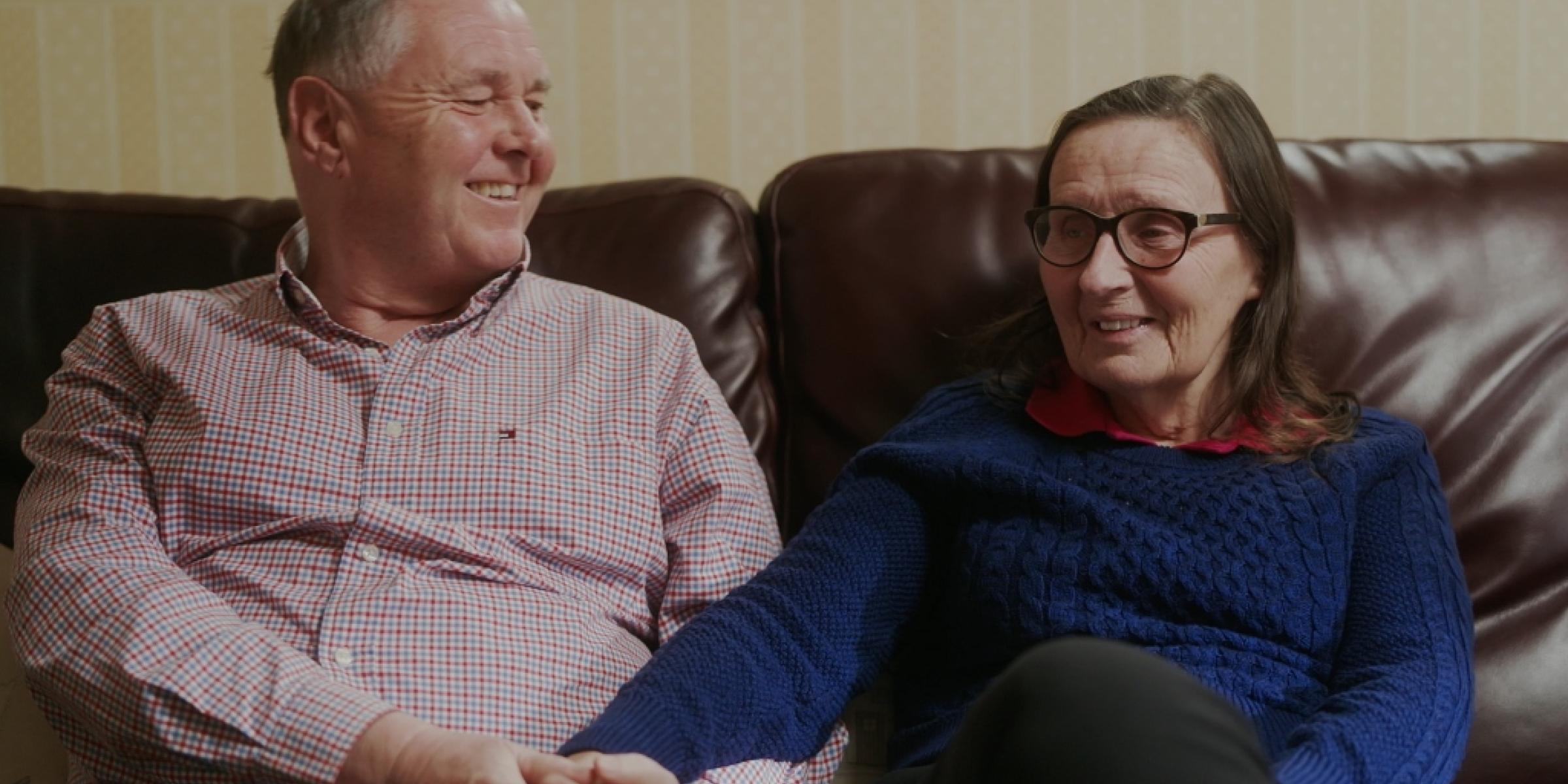 Bill and Jo sat on their couch, smiling and holding hands