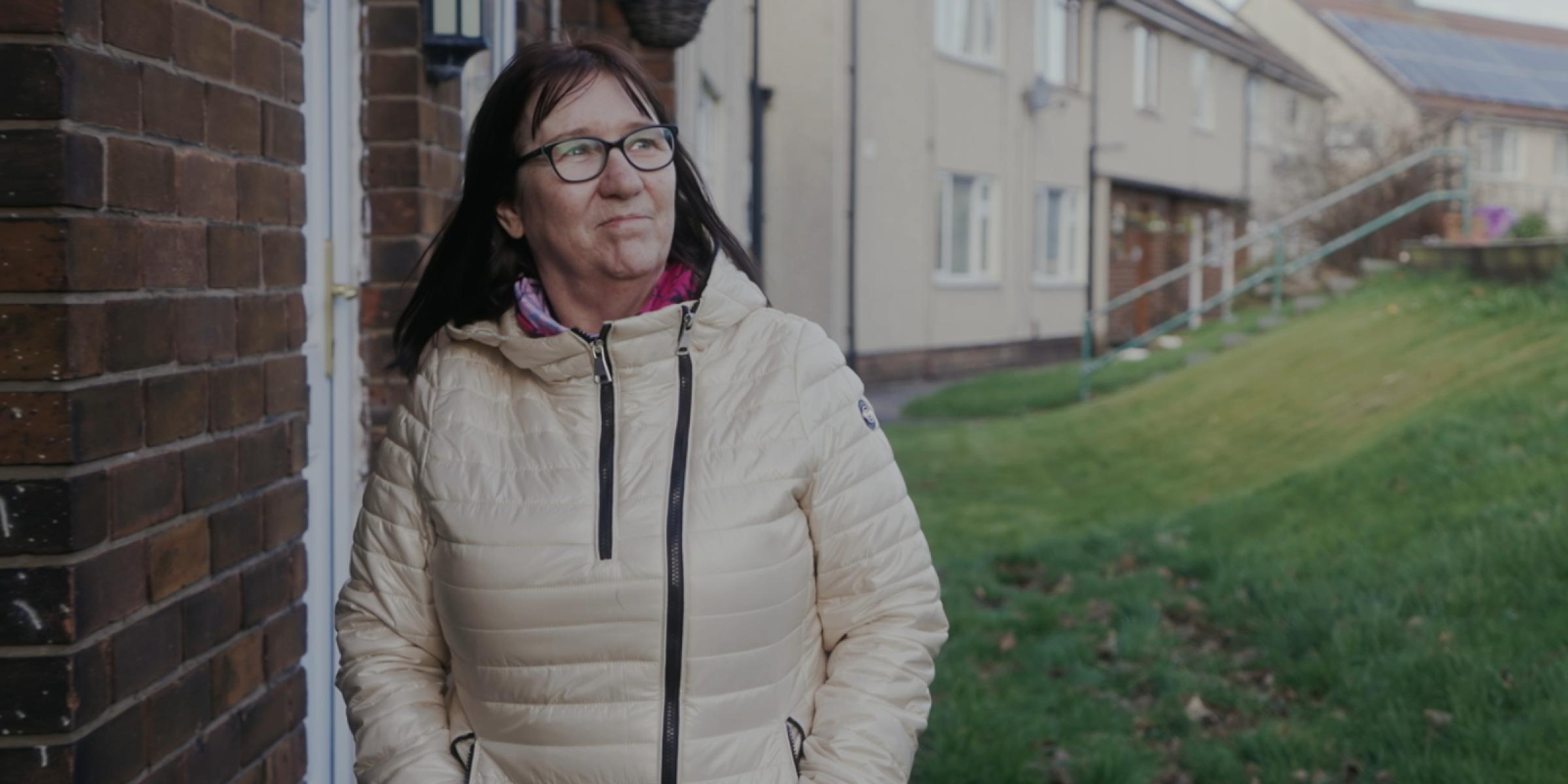 Gina walking through the neighbourhood wearing a coat with her hands in her pockets and smiling