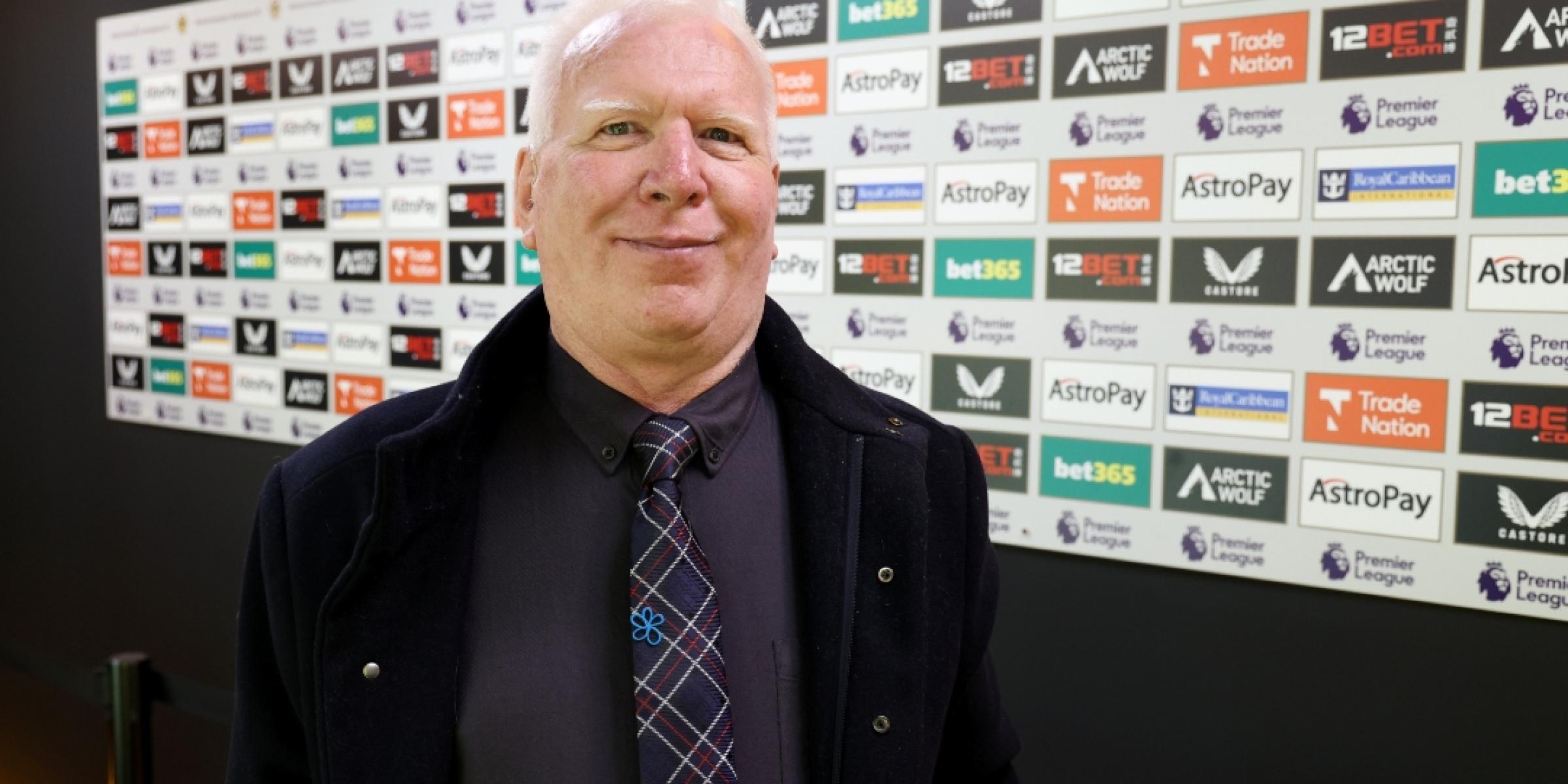 Peter Jones stood in front of a wall with advertisements on it at a football stadium, wearing an Alzheimer's Society pin badge