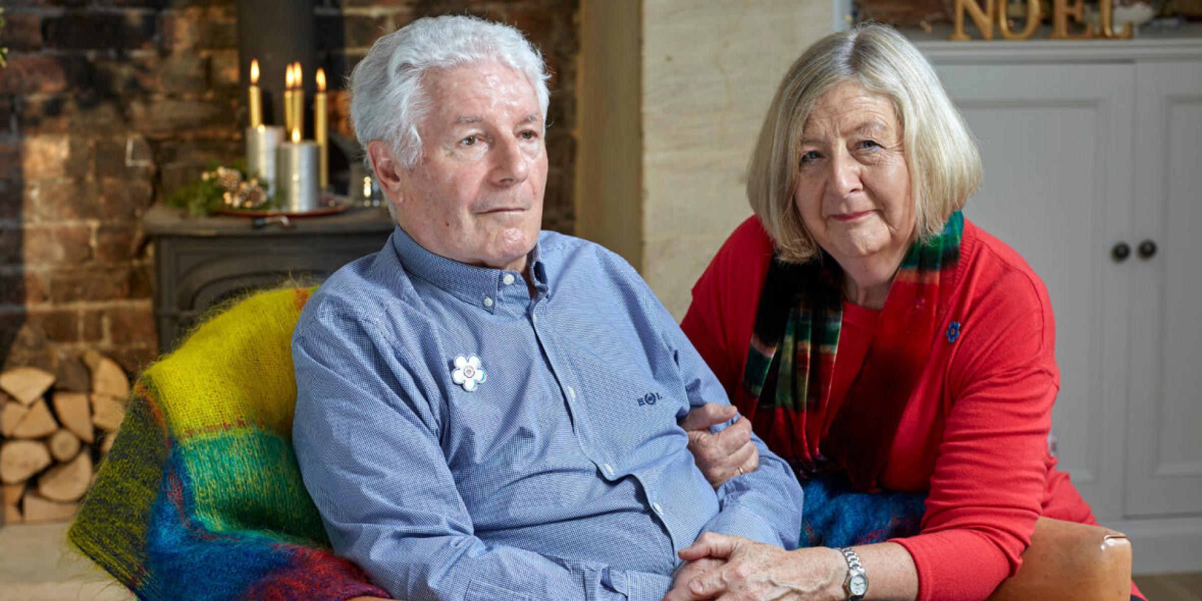 A man sat in a chair while a woman leans beside him and holds his hand