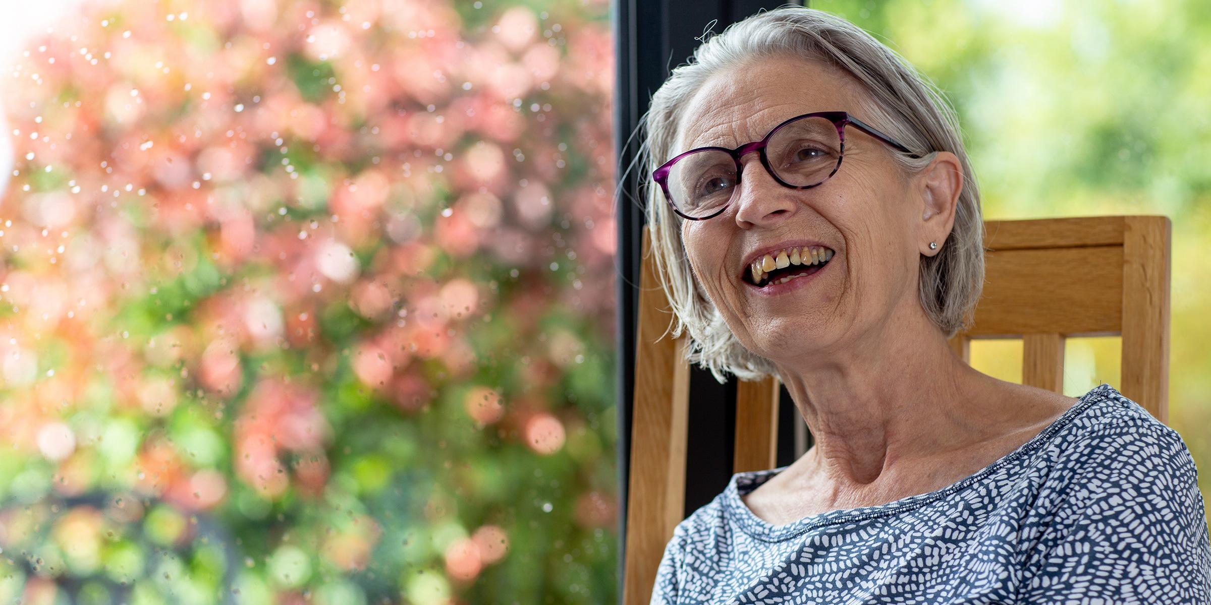 Anne Hoad sits in her home in front of the doors to her patio. She is looking to her right, smiling open-mouthed. She wears glasses and a light blue spotted tshirt. 