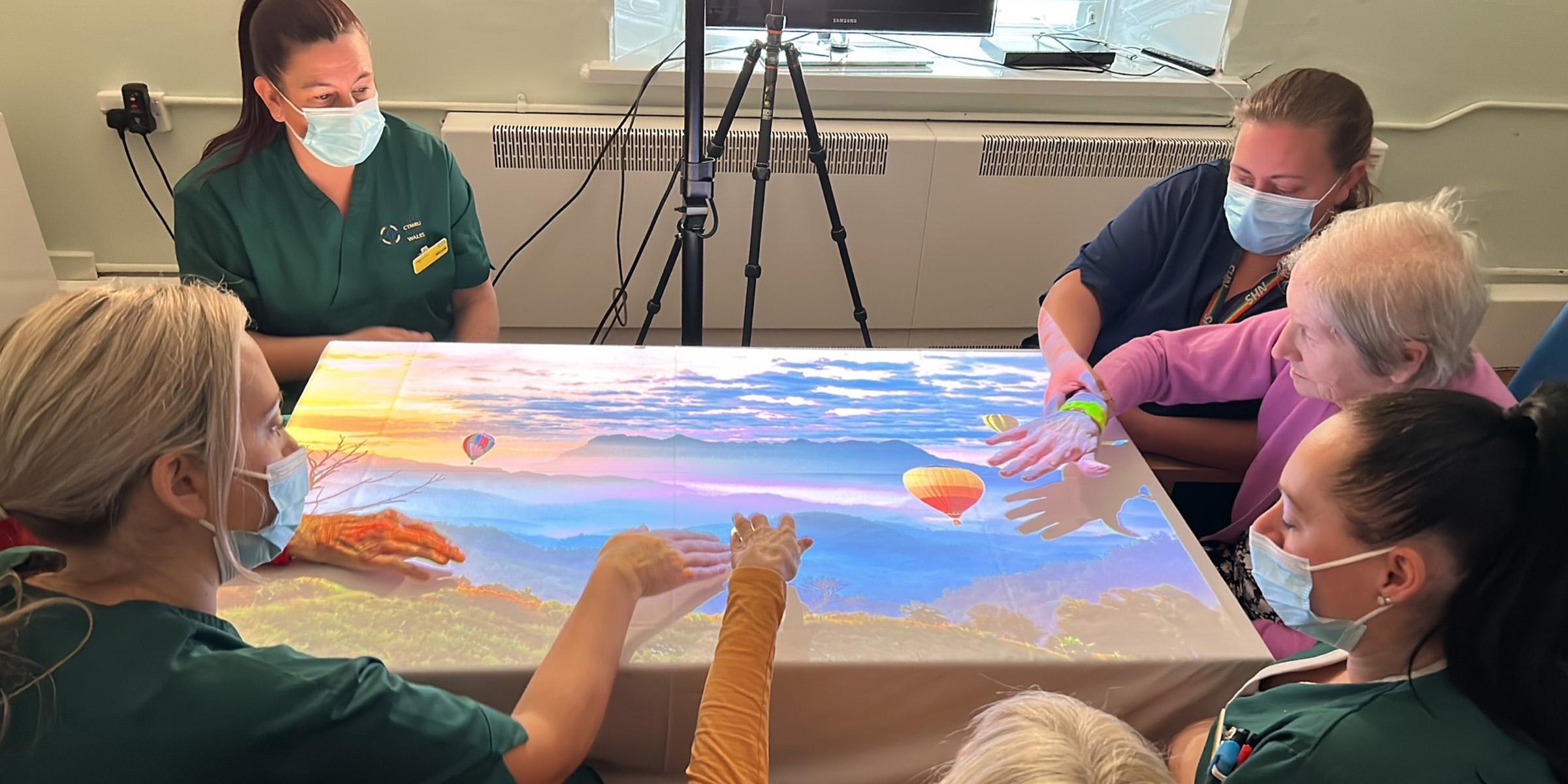 Carers and people with dementia sat around a table looking and pointing at a projected light game on the tabletop