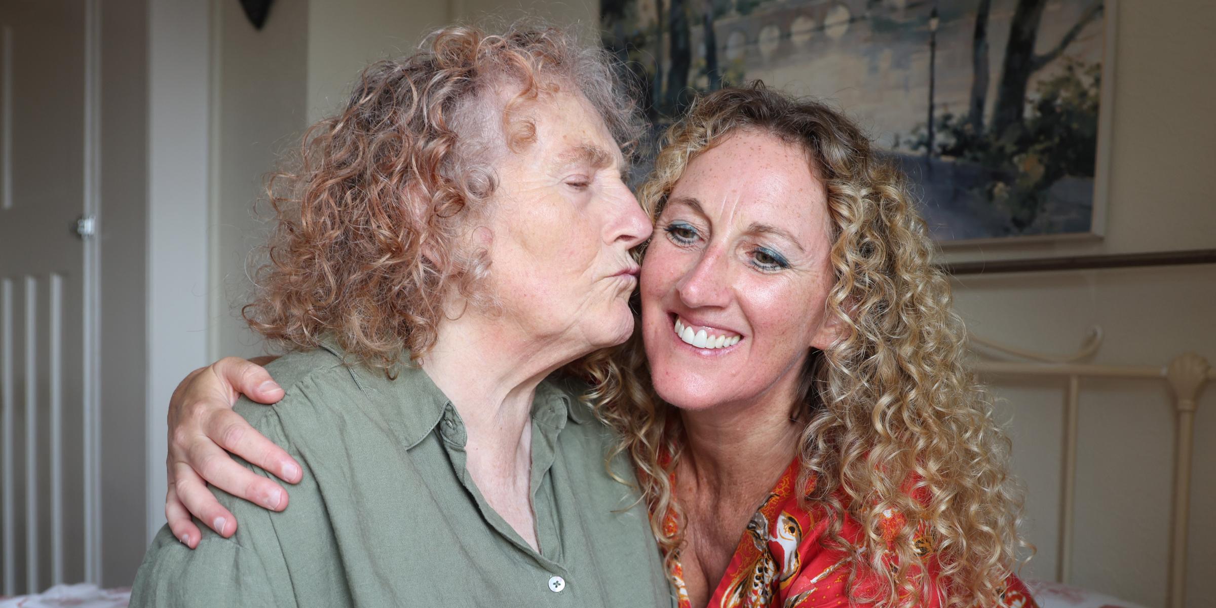 Sue gives Helen a kiss on the cheek in a bedroom