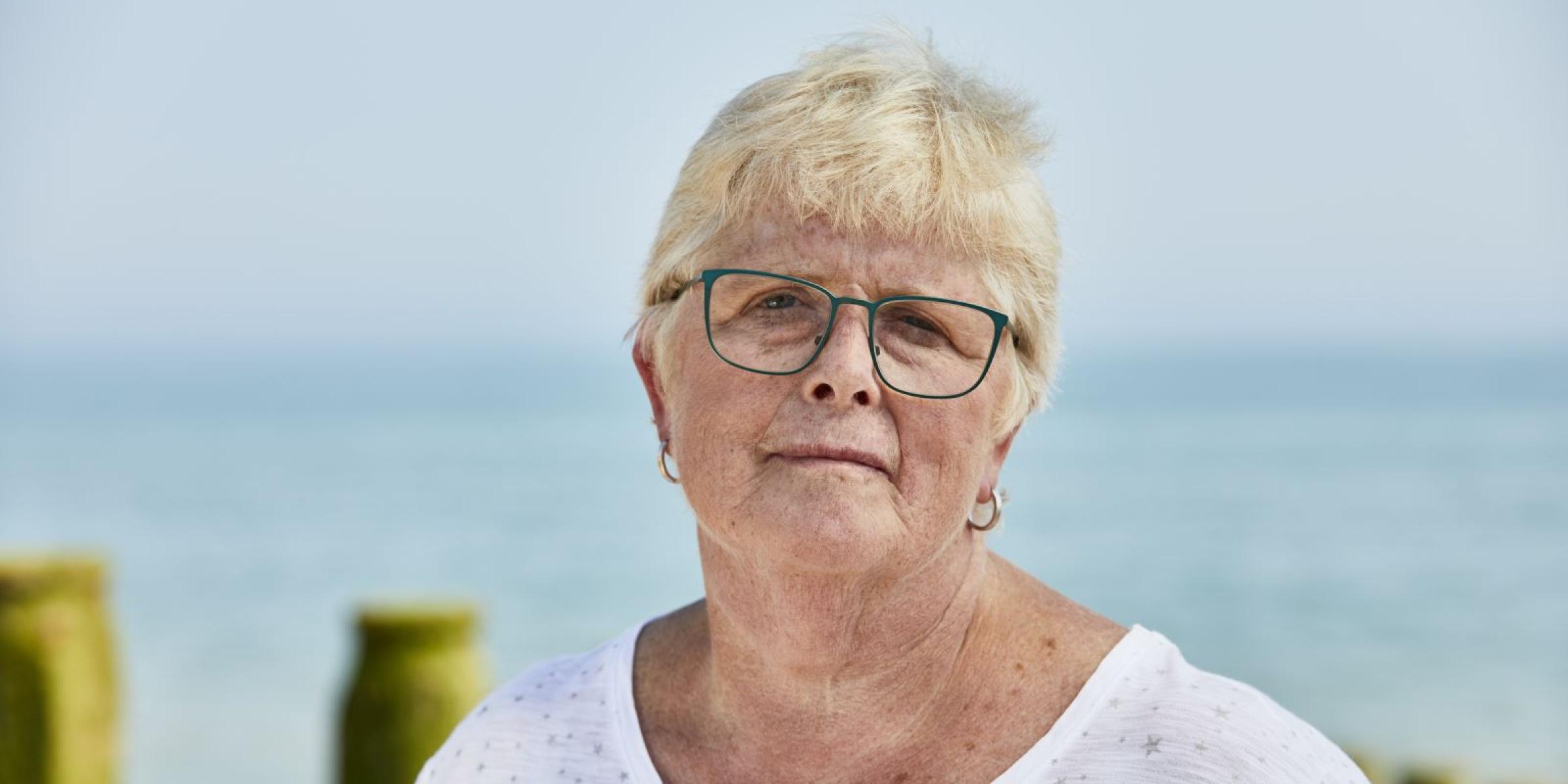 A woman with short blonde hair and glasses with the sea behind her