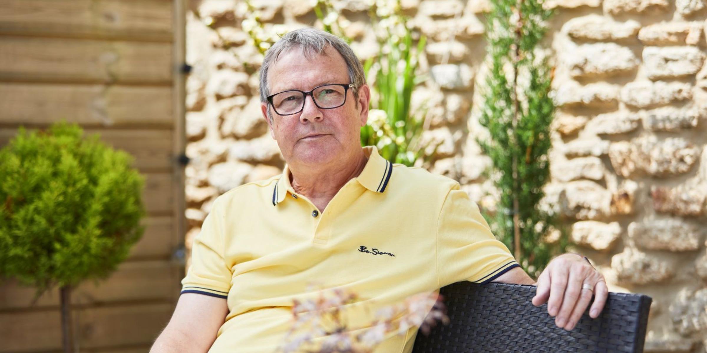 A man in a yellow shirt and glasses sits down in a garden