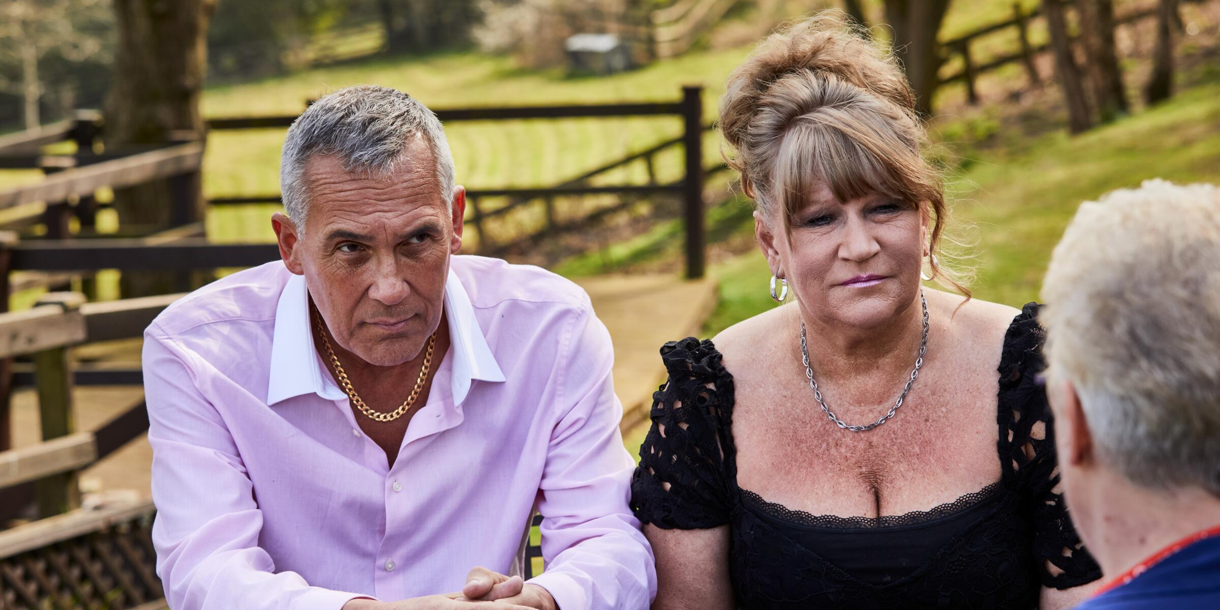 A man in a pink shirt with grey hair and a woman with in a black top with a blonde updo listening to a man with grey hair