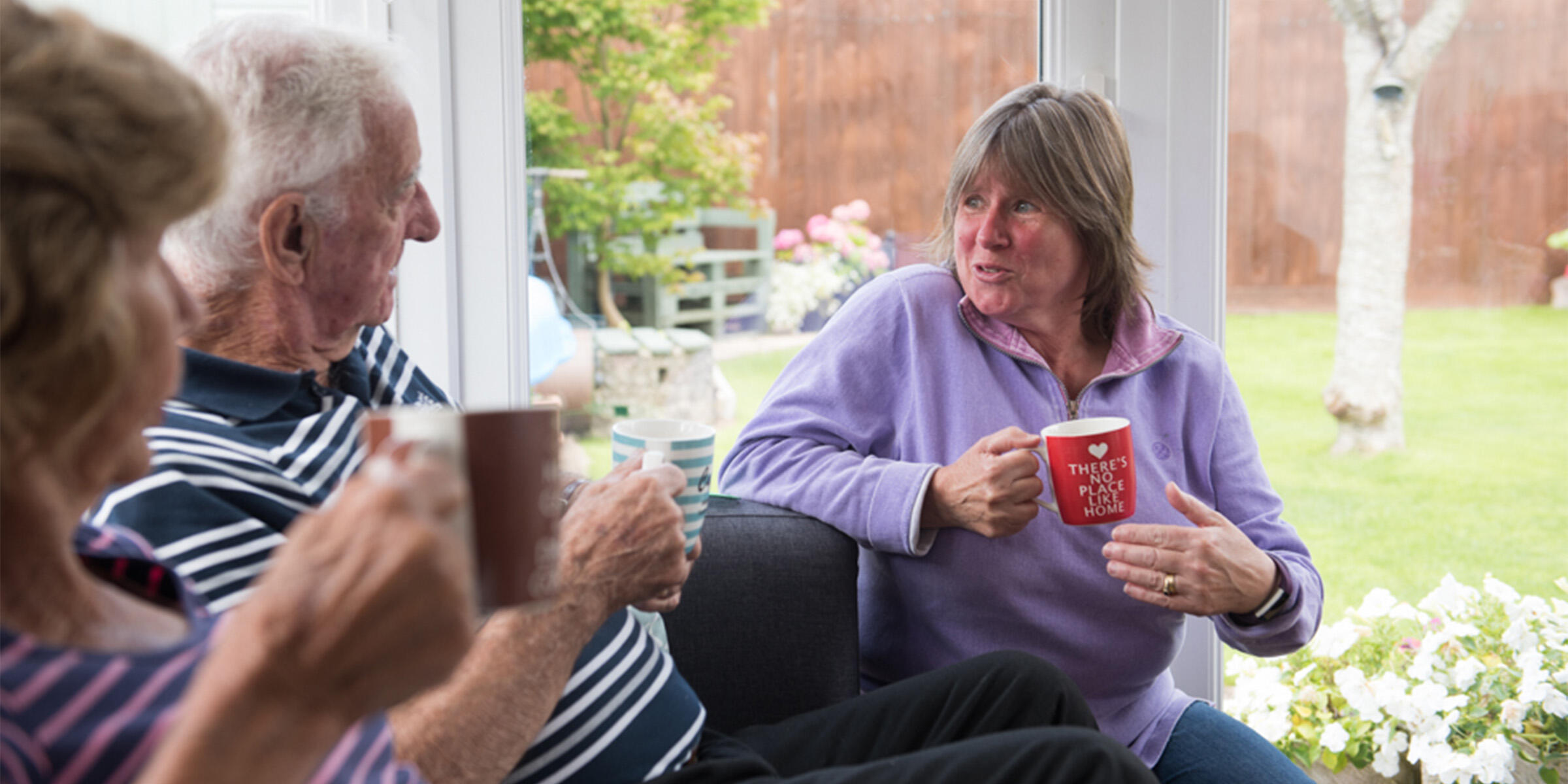 Libby is wearing a purple top and holding a cup while she talks to a man and a woman, there is a garden in the background.