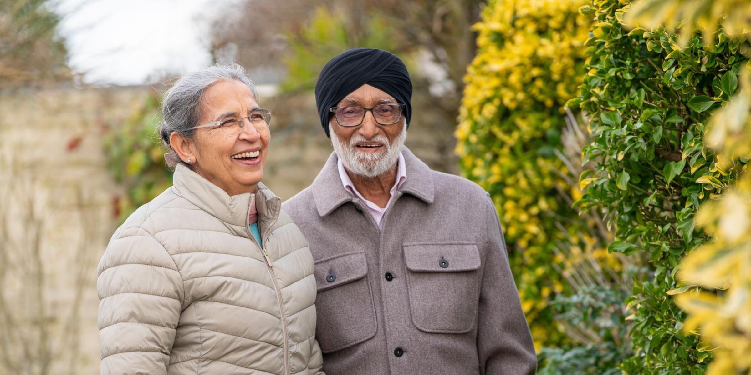 A couple wearing light coats stand in their garden smiling.