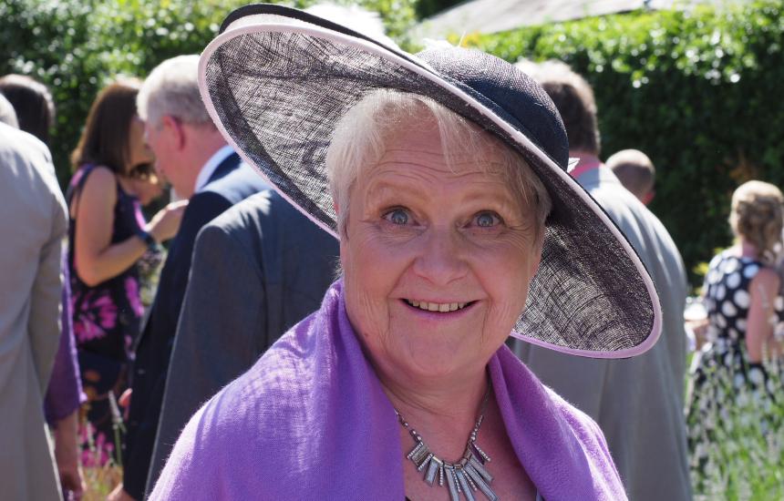 Di at a wedding wearing a large brimmed hat and a purple pashmina