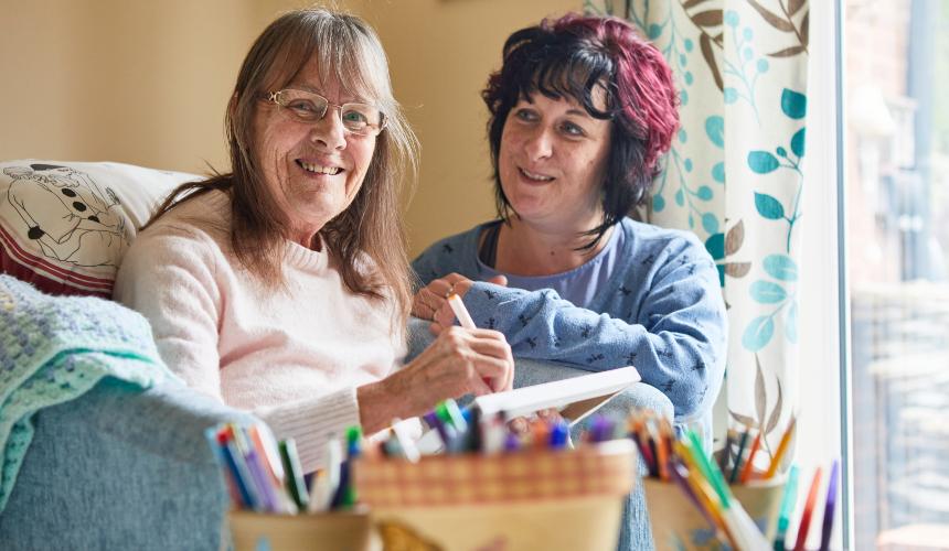 Louise sitting next to Marion in her armchair smiling with colouring stationery in the foreground