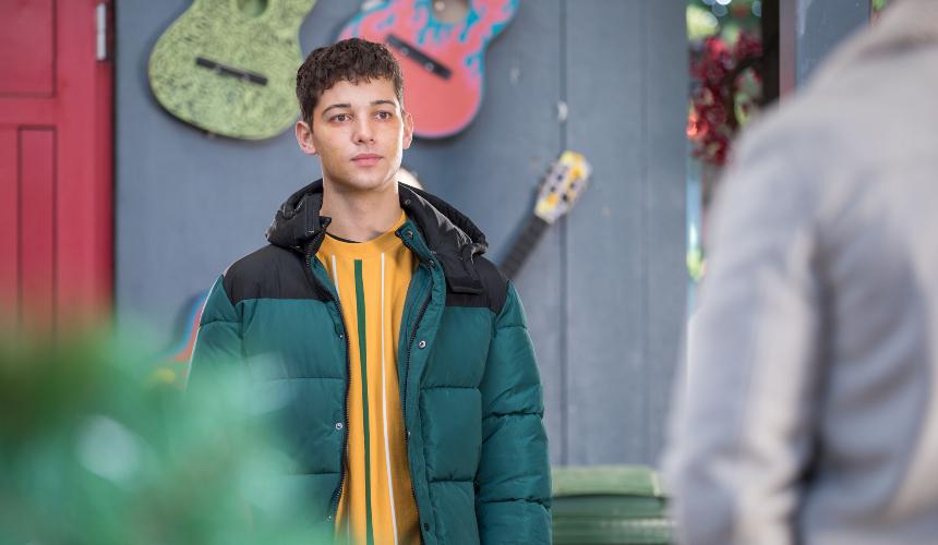 Ollie Morgan, a character from Hollyoaks, stands opposite someone with their back to the viewer with a listening expression