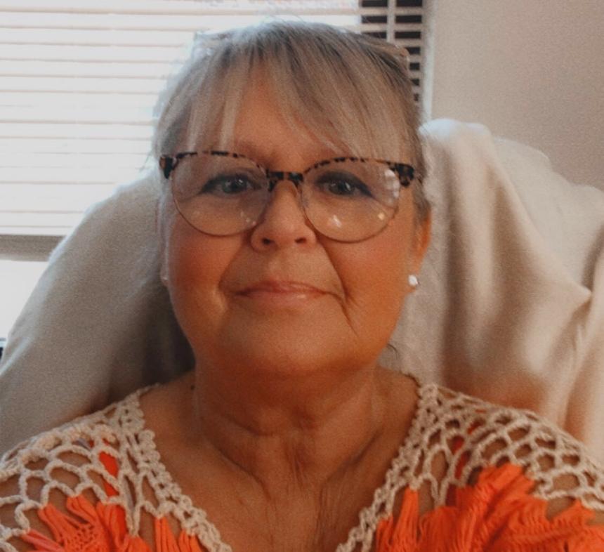 Sam is sat in her living room in an orange shirt and glasses, smiling taking a selfie