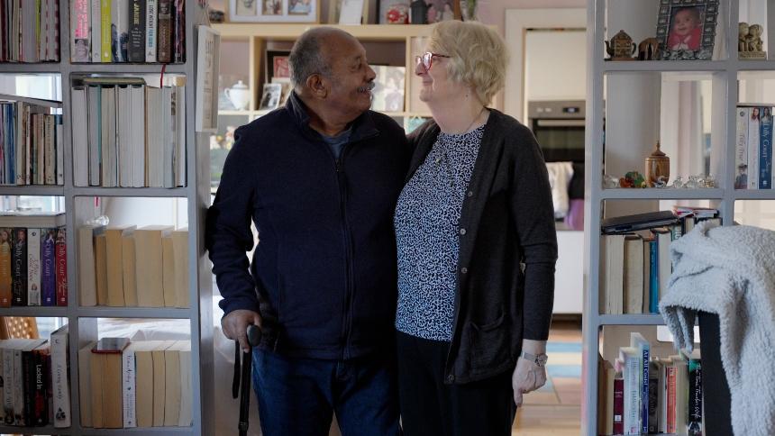 Ananga and Jo stand with their arms around each other in between two bookcases in their houses