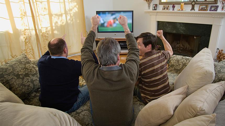 Three men celebrate a football goal in front of the TV in a living room.