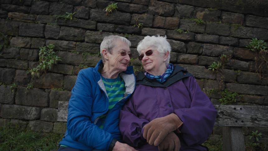 Hilary and Pauline sit closely together outside on a bench, smiling at eachother