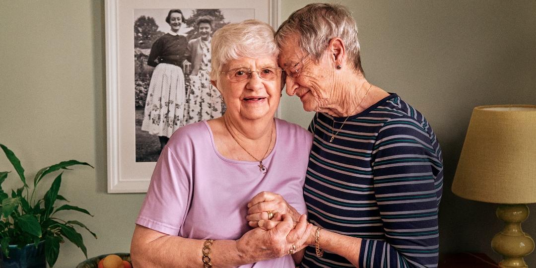Hilary and Pauline in their living room. Hilary rests her head on Pauline's shoulder and holds her hand.
