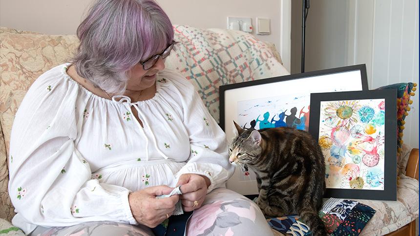 Gill Taylor sits on her sofa and looks at her cat which is sitting next to her. There are framed artworks that she has created also sitting on the sofa next to them.