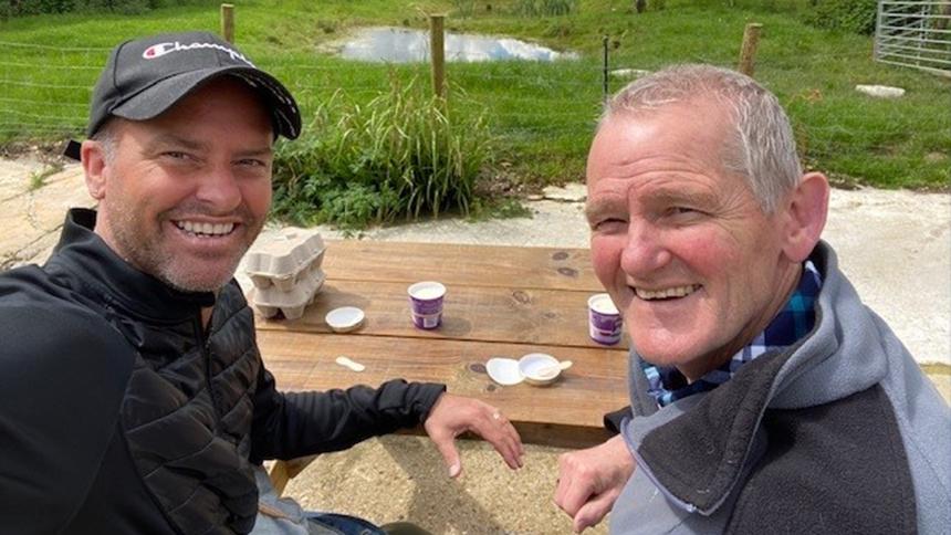 Craig and Trevor Whymark sit together on a picnic table. They're both smiling at the camera