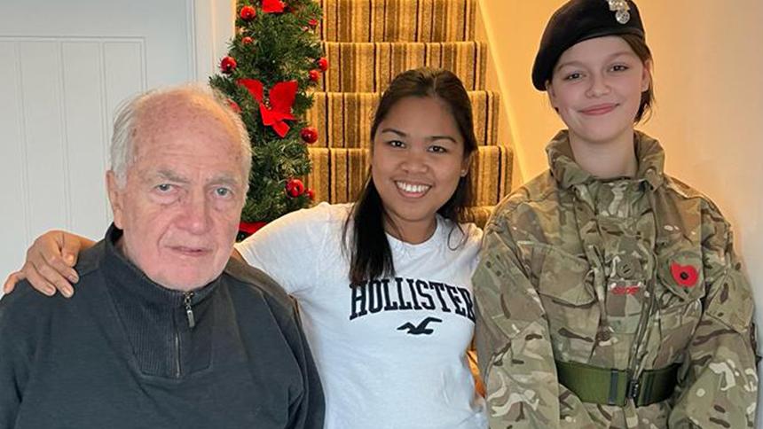 Roy and Daisy at home in front of the staircase with a family member, they have their arms round each other.