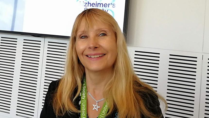 Lorraine Brown smiling, wearing a green lanyard and stood in front of a TV screen