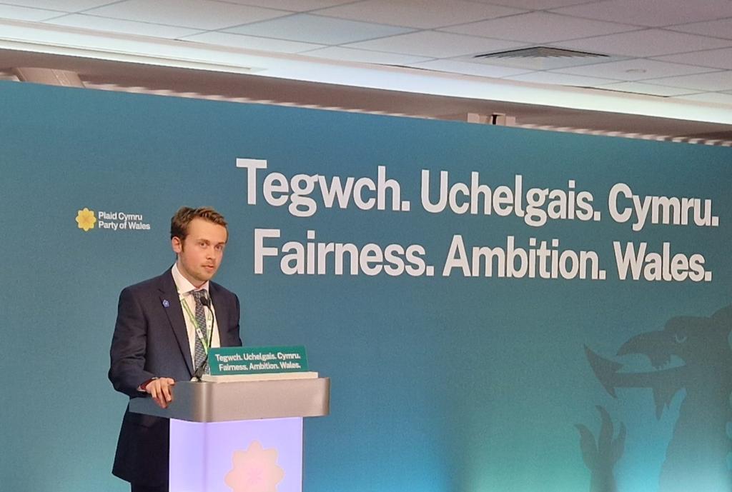 A man in a suit speaking at a podium in front of Plaid Cymru branding.