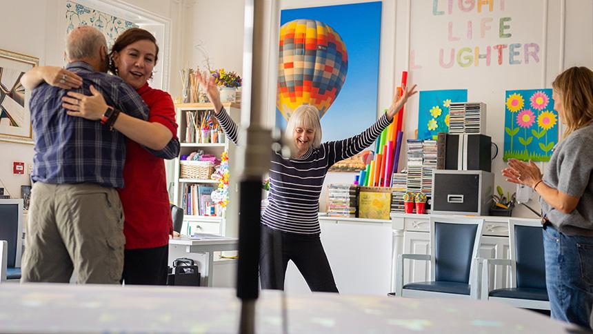 Carers and people with dementia dancing around a table in white room with bright pictures on the walls