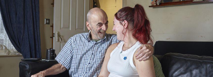 A man sits on the sofa with his arm around his daughter, laughing.