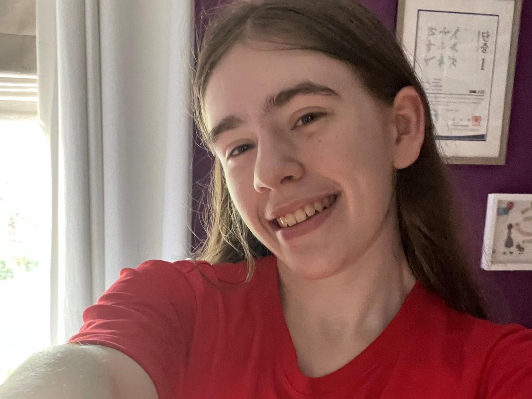 Eden has long brown hair and is wearing a red t shirt. She's taking a selfie and smiling