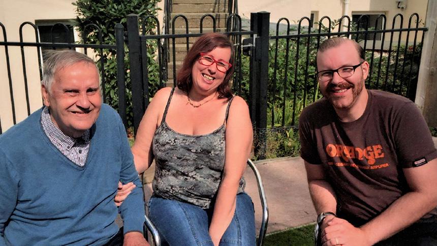 Sharon Daltrey sat in middle, wearing a t-shirt and blue jeans. To her right is her dad Brian wearing a blue jumper and to her left is her husband Chris wearing a brown t-shirt.
