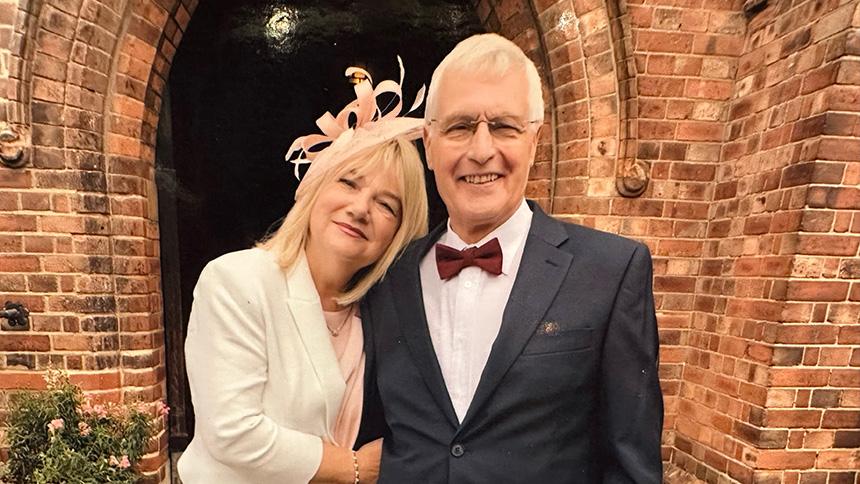 Maria and Roy smiling and stood in front of a red brick church at a wedding, Maria is wearing a cream and pink outfit and Roy is wearing a dark suit and bowtie