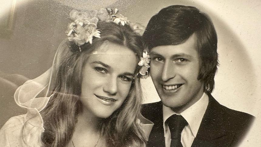 A black and white wedding photo of Maria and Roy smiling, Maria is wearing a flowery headdress, Roy wearing a dark suit and tie.