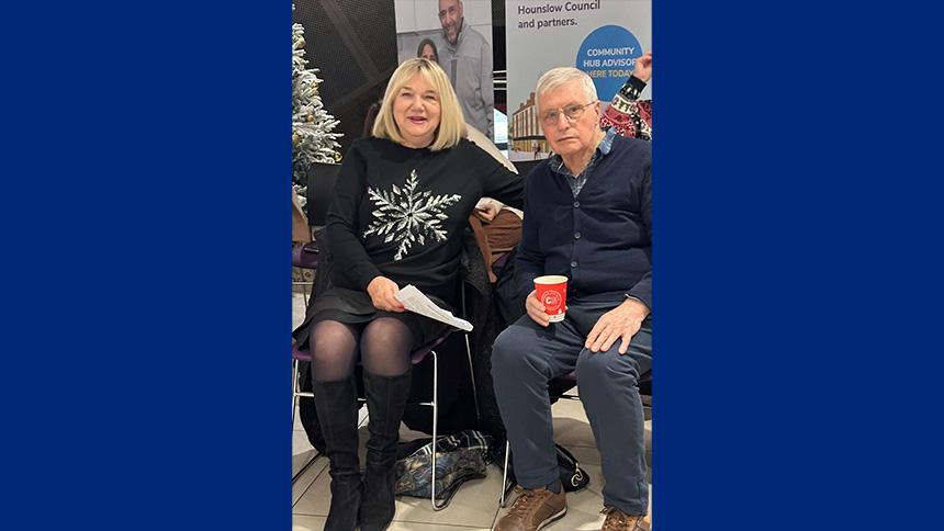 Maria and Roy sat down at a Singing for the Brain Christmas meeting, Maria is wearing a black Chrstmas jumper, Roy is holding a cup of tea and wearing a dark-coloured jacket.
