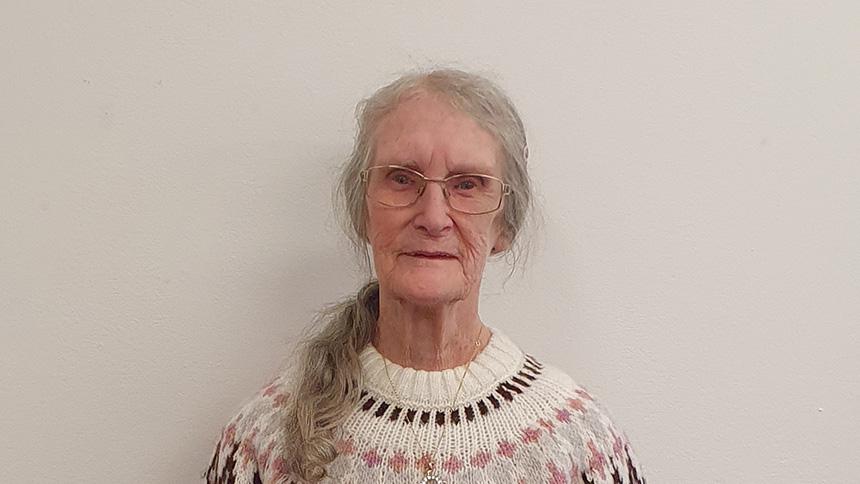 Margaret wearing a white and pink jumper, she has long grey hair and wears glasses