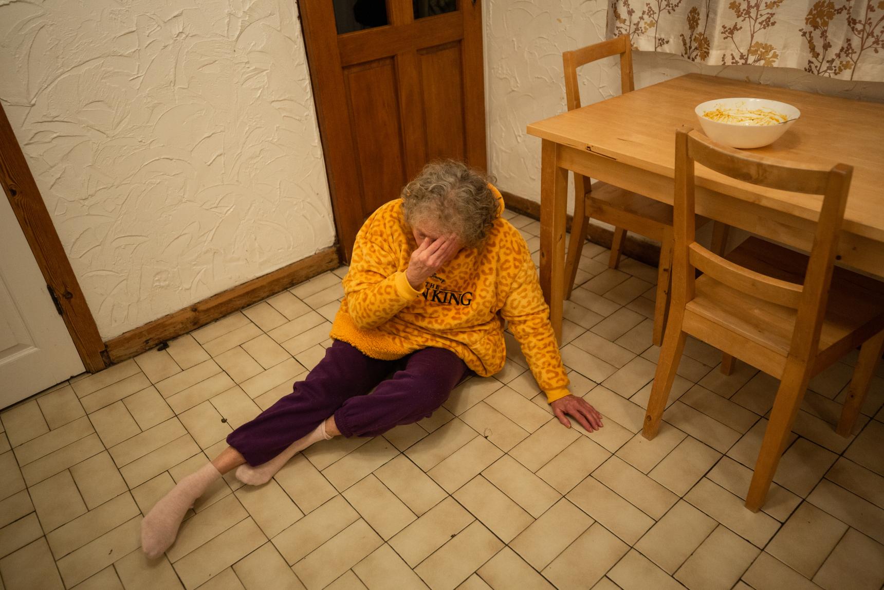An elderly lady on a kitchen floor with her head in her hand
