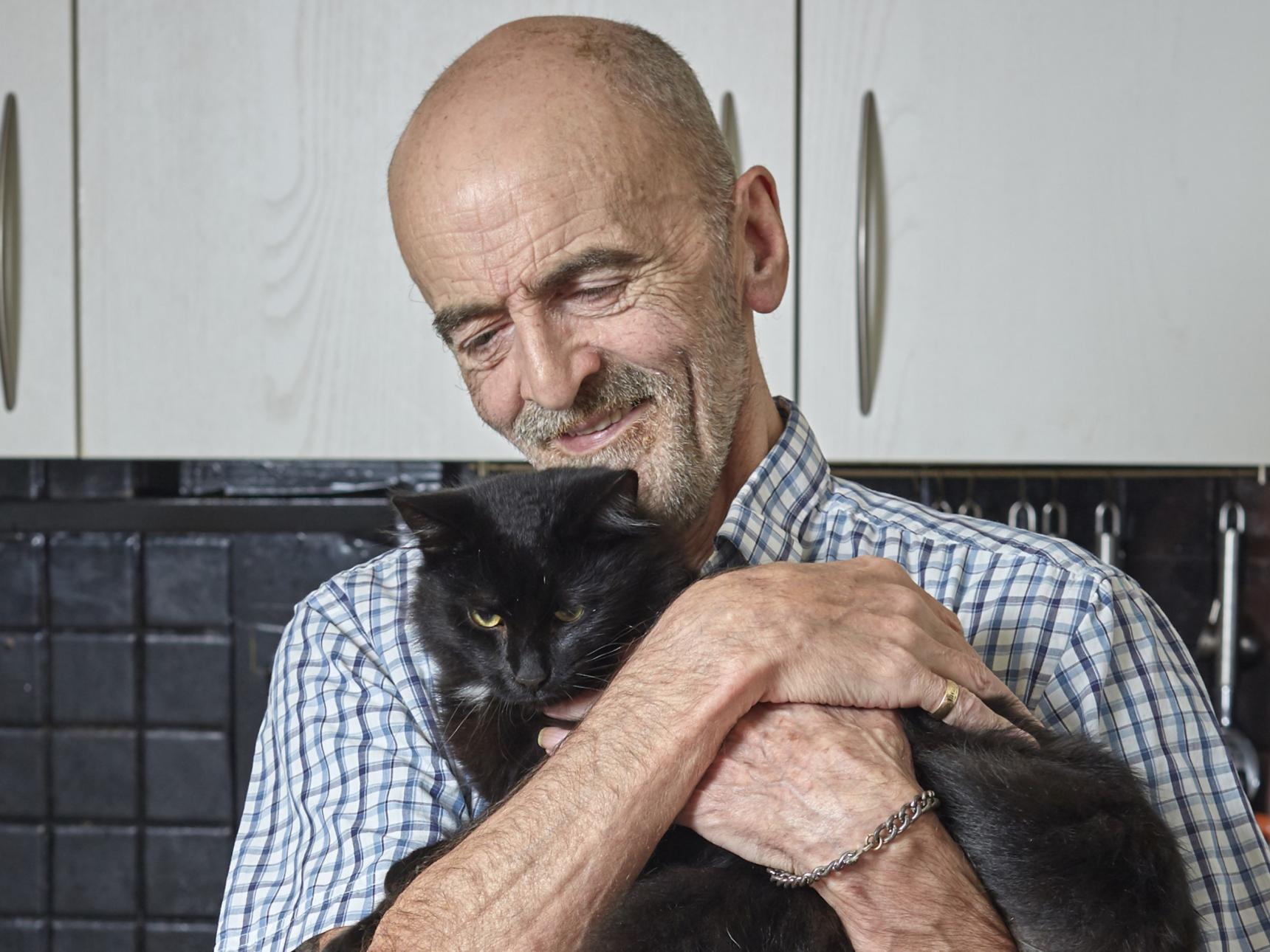 A bald man with a blue checked shirt smiles while holding a black cat