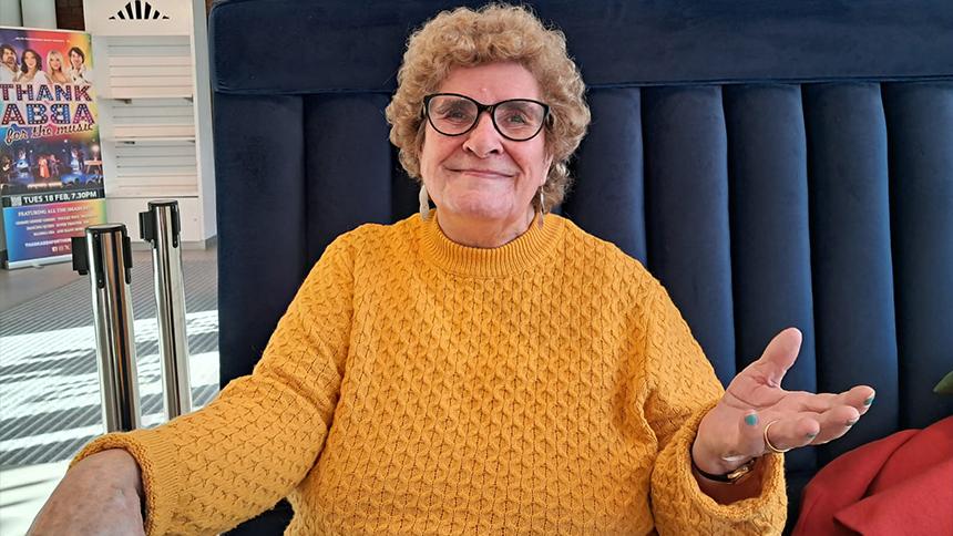 Maureen is wearing a bright yellow jumper and sat on blue seating