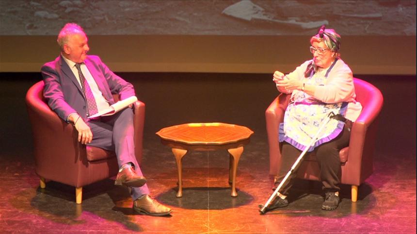 Maureen sat on stage on a brown leather chair, wearing a pinny talking with Peter who is also sat down and wearing a grey suit.