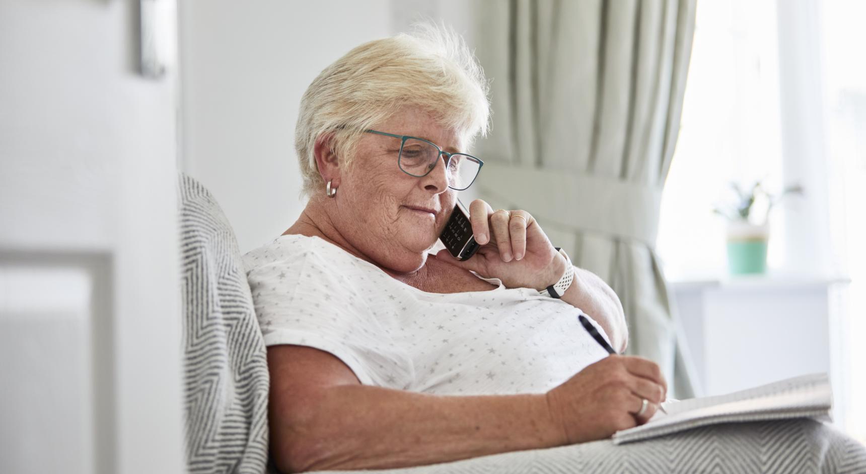 A woman sits on her sofa writing notes while listening on the phone.