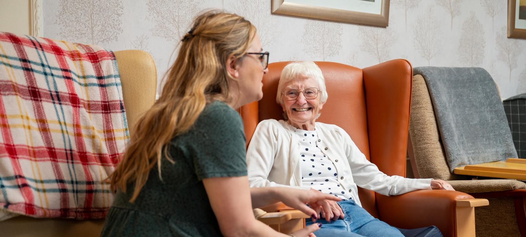 nAn older woman sits in an armchair smiling and listening to a younger woman