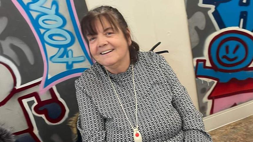 Irene is sat down in front of a wall of street art, she has brown hair and is wearing a blue top