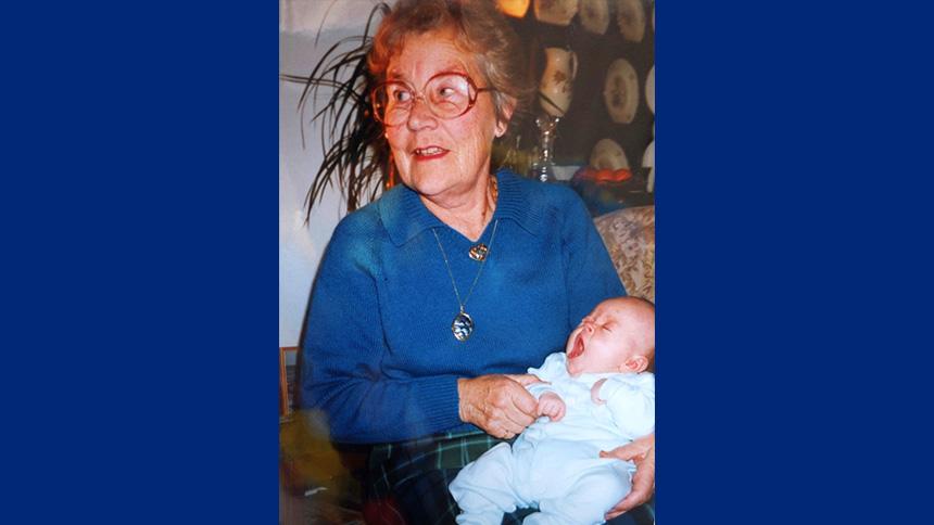 Baby Kerri in a white babygrow, being held by her nan who has glasses and is wearing a blue blouse