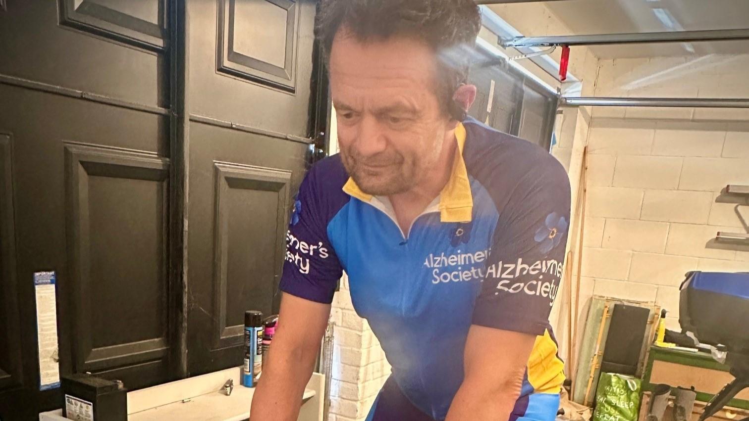 A man in Alzheimer's Society cycling shorts and t shirt on an exercise bike in a garage