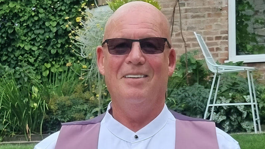 John is sat in a garden, wearing sunglasses, a white shirt and pink waistcoat.